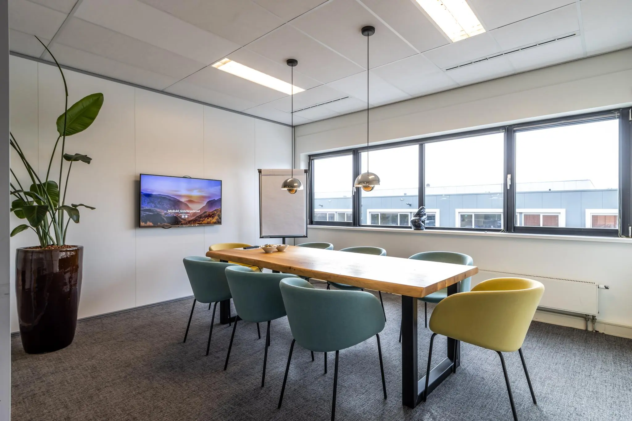 Vergaderruimte aan de Lage Biezenweg met modern interieur, grote houten tafel, kleurrijke stoelen en een presentatiescherm.